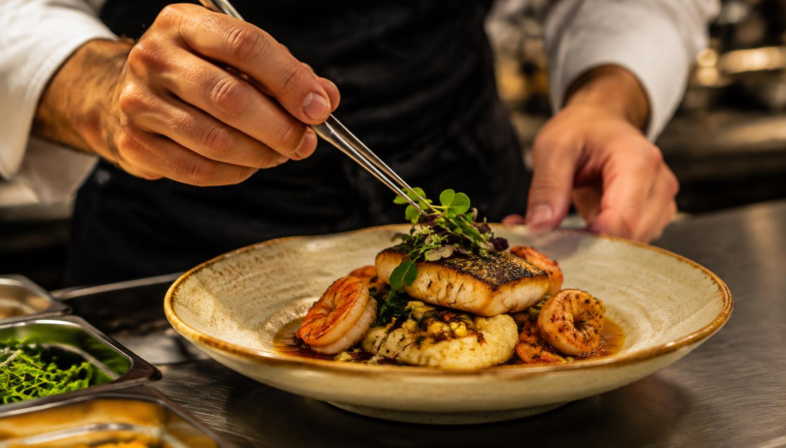 A chef's hands plating fish and shrimp with tweezers